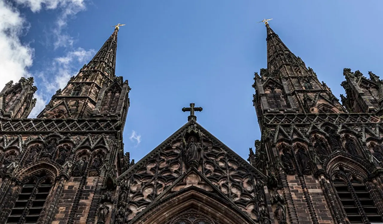 Two tall Gothic cathedral spires with ornate stonework rise against a blue sky with white clouds.