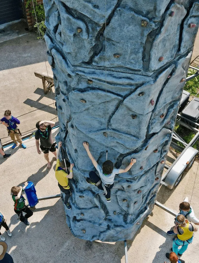 Children climbing a tall blue artificial rock climbing wall with handholds, belayed by instructors on the ground below.