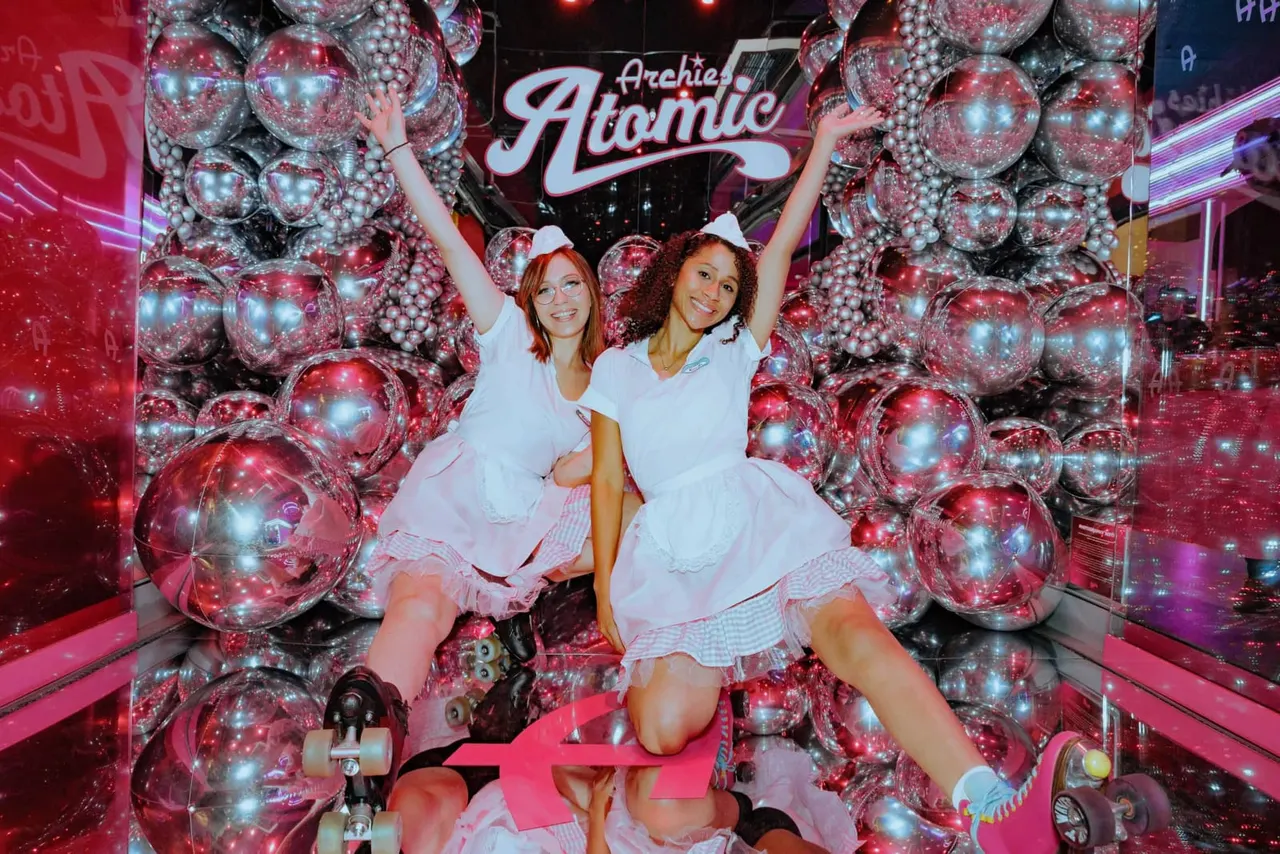 Two women in white outfits posing with raised arms in front of illuminated disco balls at a retro-themed venue.
