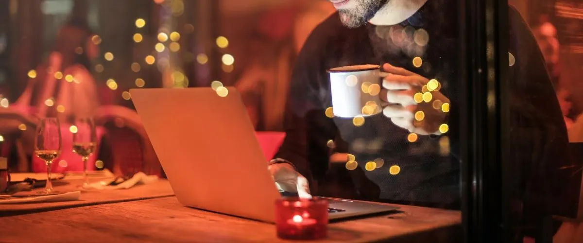 Man holding a white mug at a table with a laptop, surrounded by warm golden lights and festive decorations.