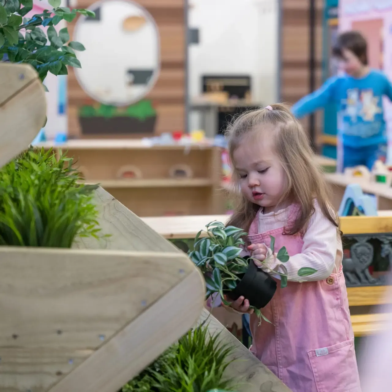 A young girl in a pink apron holds a potted plant in an indoor play space with wooden shelving and other children in the background.