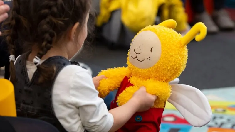 Child holding a yellow soft toy character with antennae during an indoor activity session.