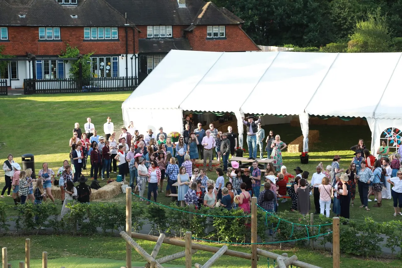 Large outdoor event with many guests gathered around white marquees on a lawn beside a brick building.