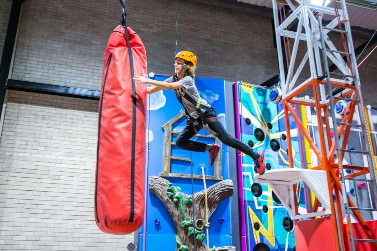 Person in safety helmet suspended mid-leap towards a red punching bag at an indoor climbing or activity centre.