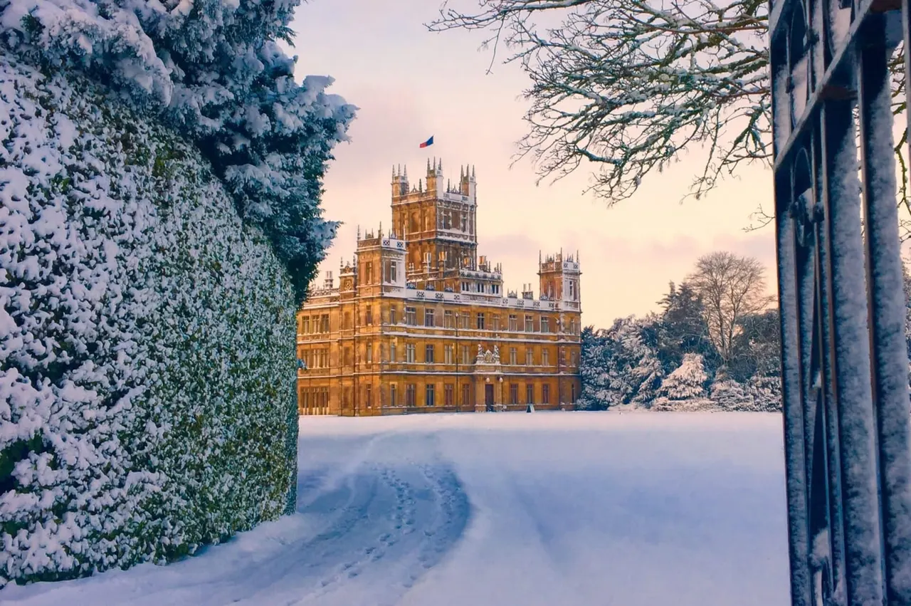 Snow-covered grounds with frost-laden hedges framing a grand Victorian castle in winter sunshine.