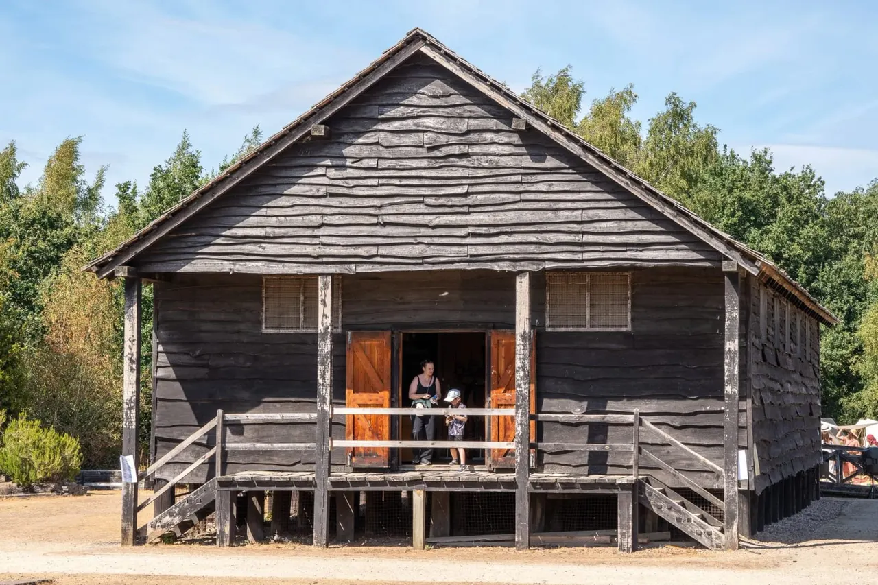 An adult and child stand on the verandah of a reconstructed dark wooden Roman fort building with a pitched roof, surrounded by trees.
