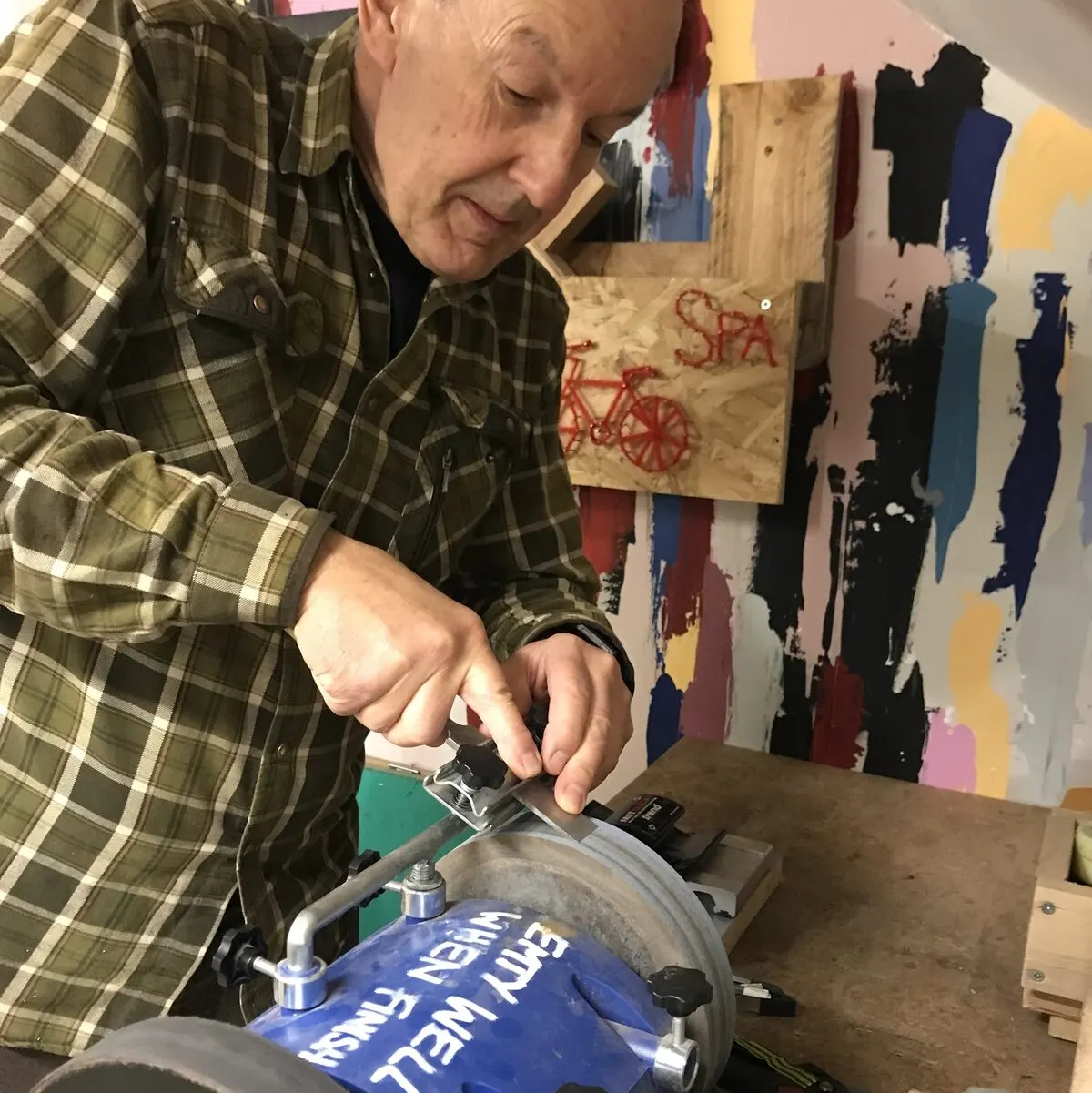 A person in a green plaid shirt uses a circular saw to cut a blue piece of material in a workshop.