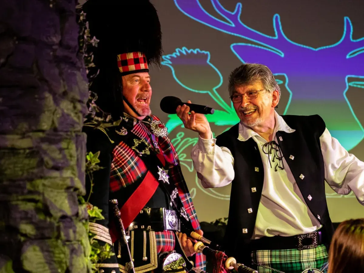 Two men in Scottish traditional dress perform on stage, one singing into a microphone and wearing a tall black fur hat, with neon antler graphics projected behind them.