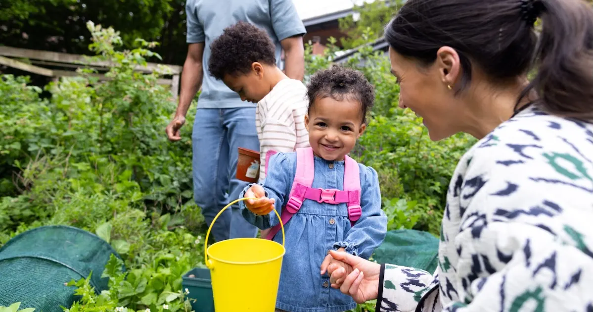 Young children and adults picking vegetables in a garden, with a child holding a yellow bucket and a harvested tomato.