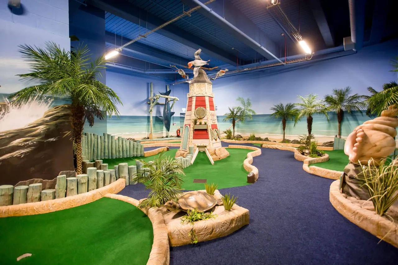 Indoor adventure golf course with tropical beach-themed decorations, including palm trees, a red and white lighthouse, and a seaside backdrop.