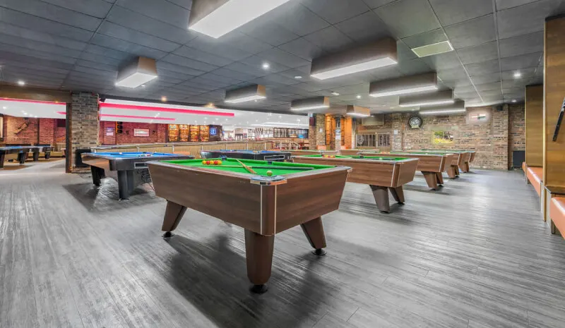 Multiple pool tables with green felt surfaces in a spacious indoor games room with brick walls and bright lighting.