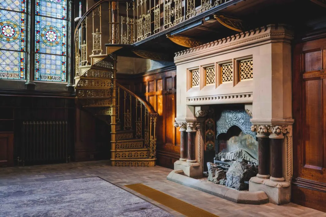 Ornate interior of a historic building featuring a grand spiral staircase, decorative fireplace, and stained glass windows.
