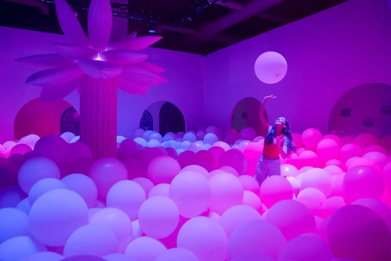 Child standing in a ball pit filled with illuminated pink and blue balls under neon purple lighting.