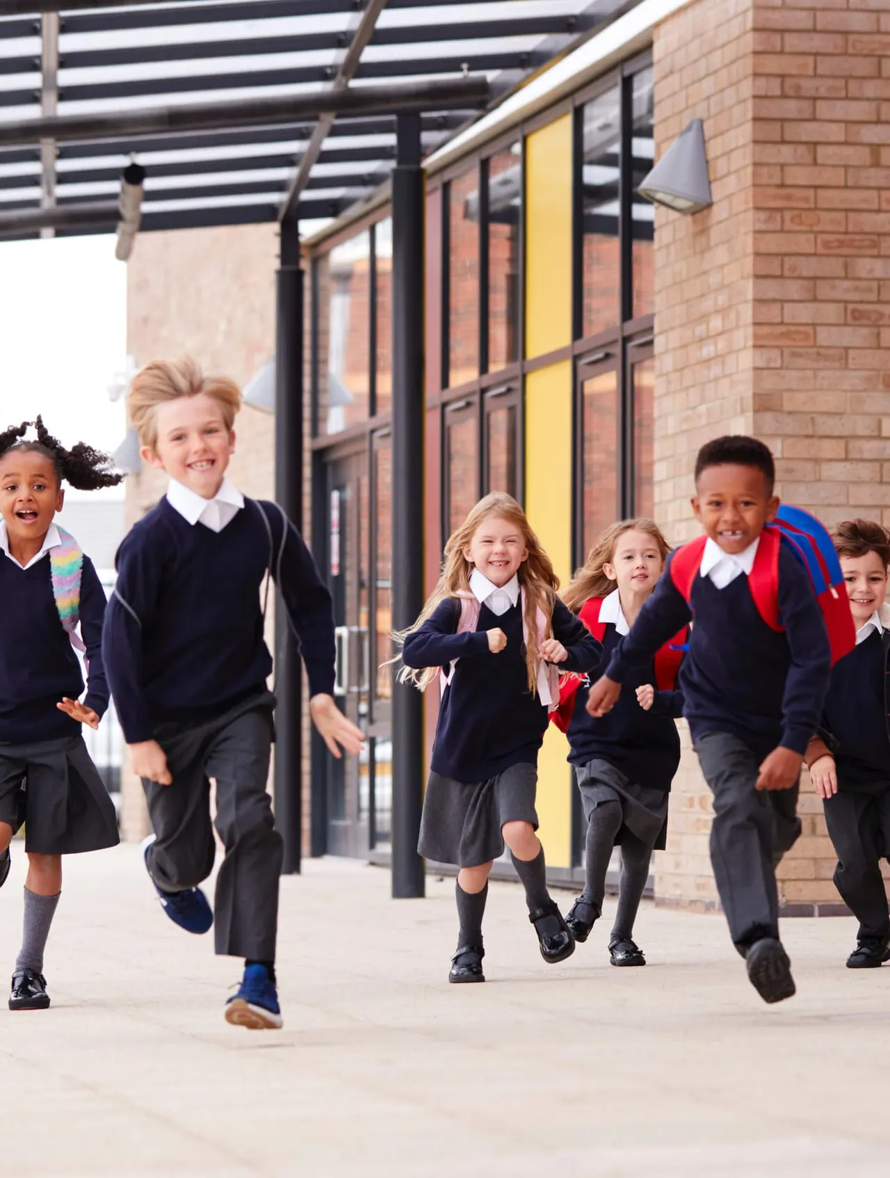 Group of primary school children in uniforms running together outside a modern brick building with yellow doors.