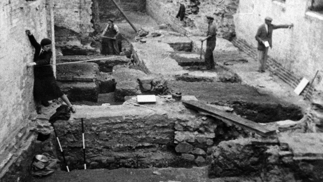 Black and white photograph of an archaeological excavation site with stone foundations and several workers examining the dig.