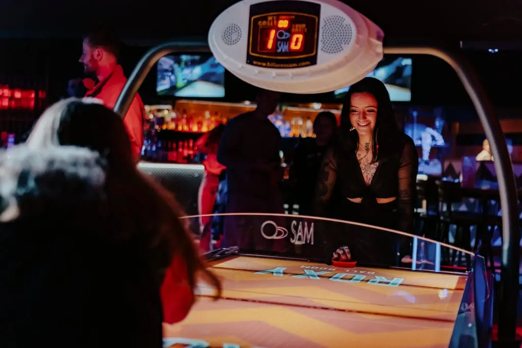 People playing air hockey at an indoor entertainment venue lit by neon lighting.
