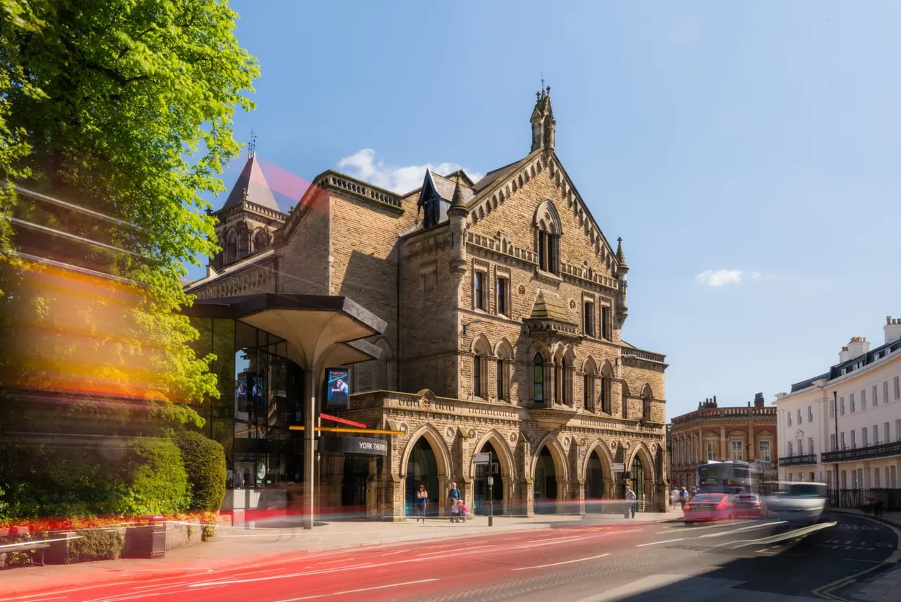 Historic stone theatre building with Gothic architecture and modern entrance canopy on a sunny day, with traffic and pedestrians outside.