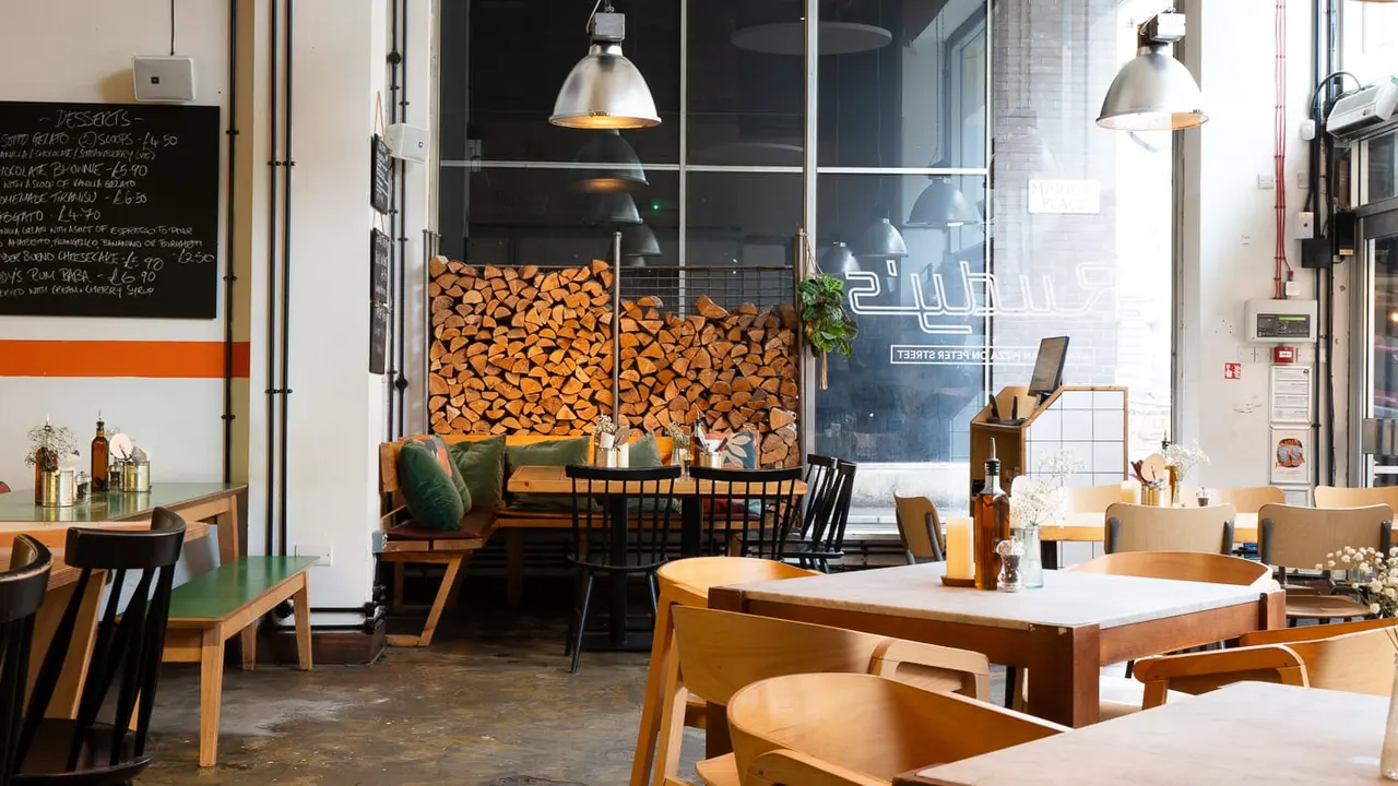 Modern restaurant interior with wooden tables, chairs, pendant lighting, and stacked firewood visible through large windows.