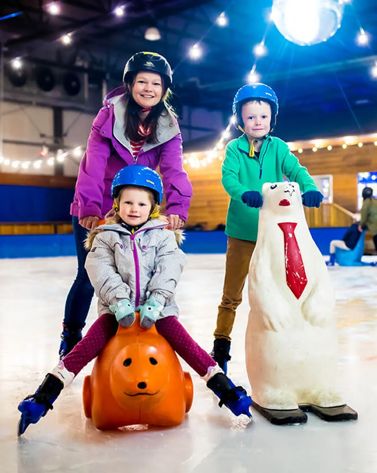 Three children on an ice rink with novelty characters, one sitting on an orange pumpkin head prop and another holding a white snowman figure.