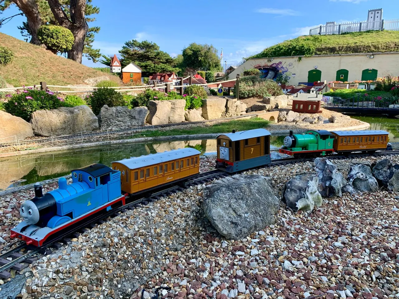 Miniature railway train with blue engine and brown carriages running on track through model village landscape with pond and buildings.