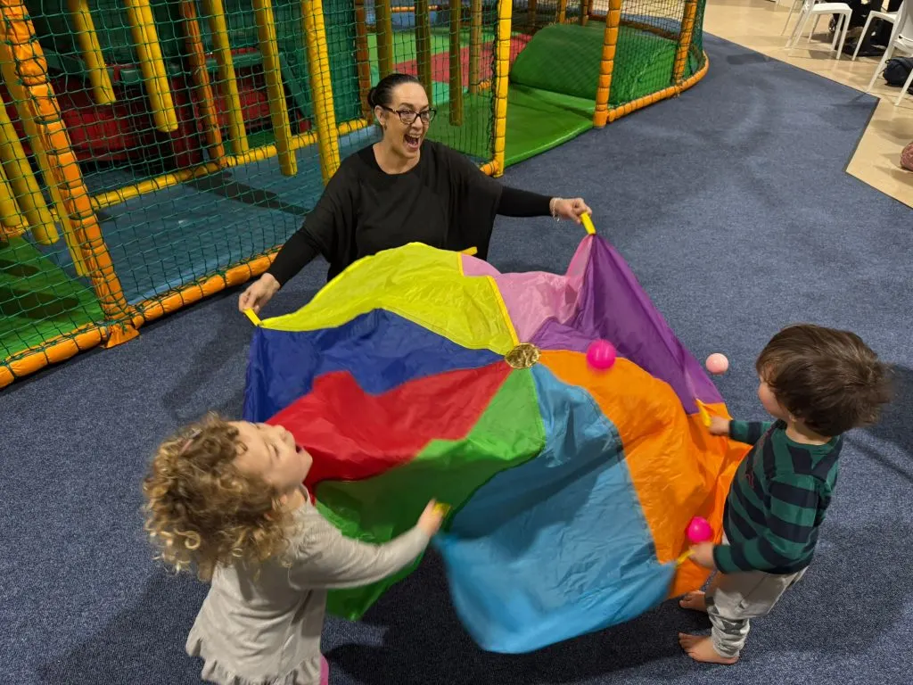 An adult and two young children play with a colourful parachute indoors next to a soft play structure.