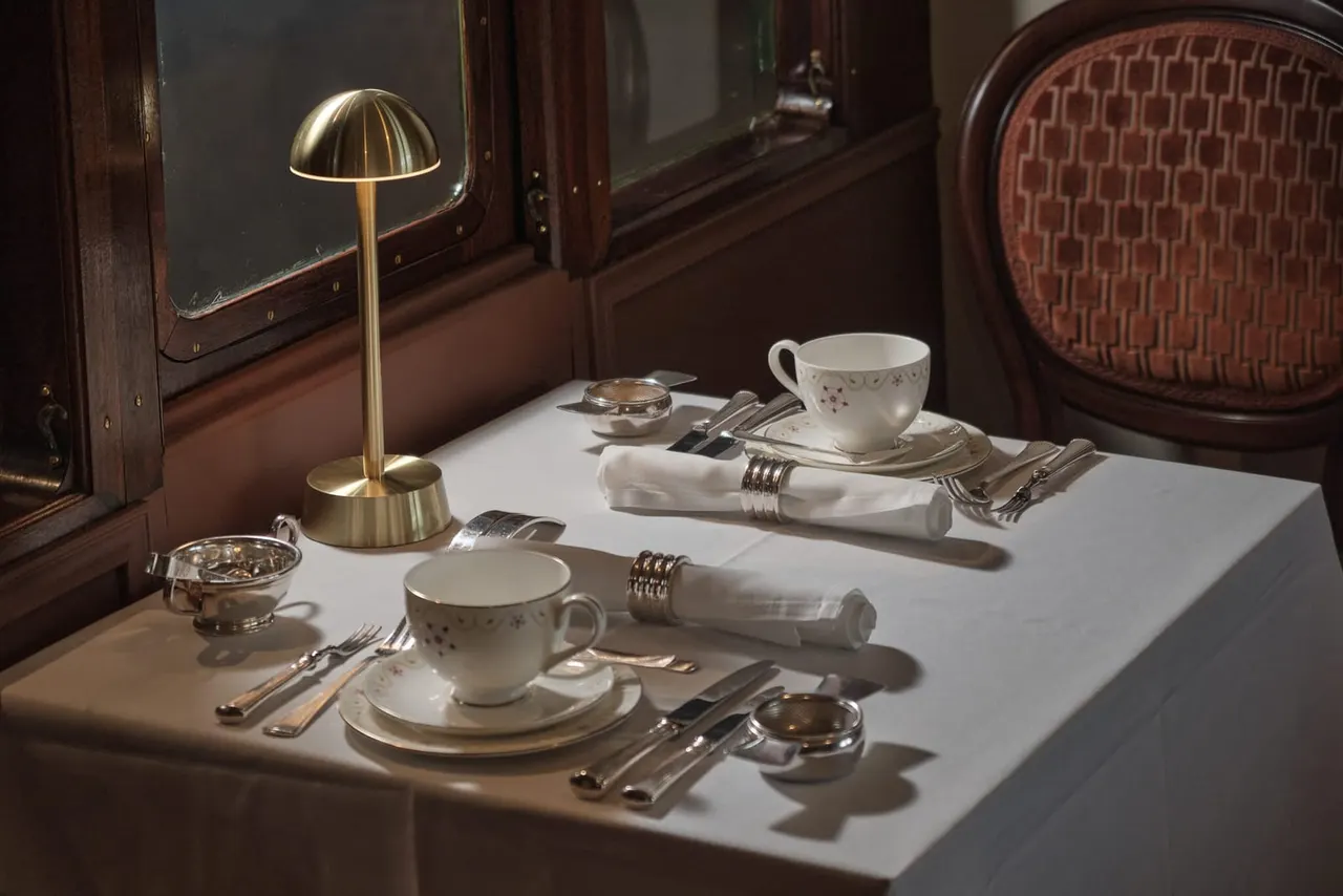 A formally laid dining table inside a vintage railway carriage with fine china, cutlery, napkins and a brass lamp.