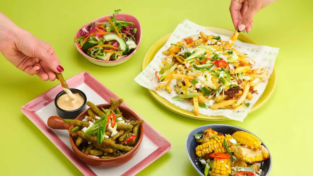 Hands dipping into dipping sauce beside loaded fries, nachos, salad and corn dishes on a bright lime green background.