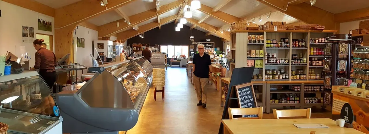 Interior of a farm shop with wooden beams, display counters, shelving stocked with products, and customers browsing.