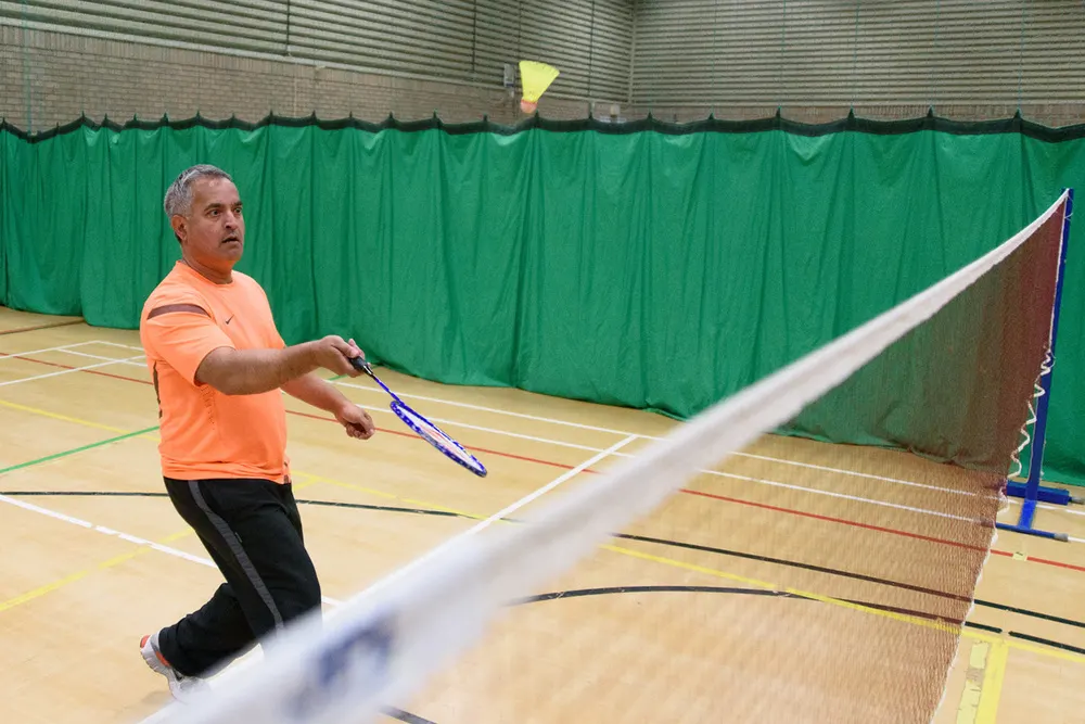 Man in orange shirt playing badminton indoors on wooden court with green backdrop.
