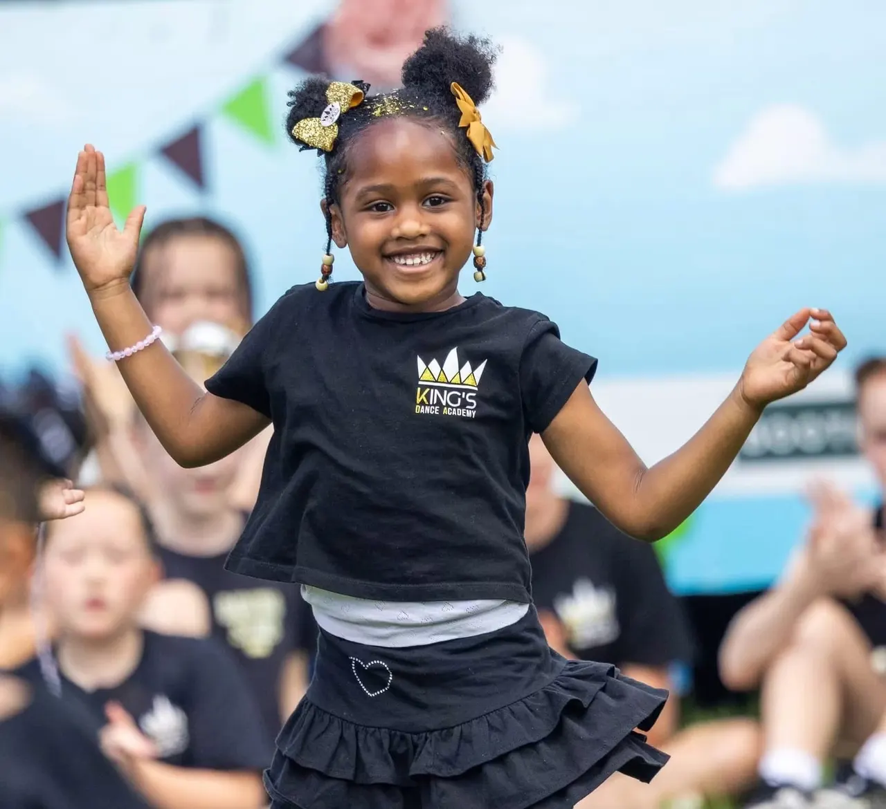 Young child performing a dance move outdoors with arms raised, wearing a black King's Dance Academy t-shirt and hair accessories, smiling at the camera.