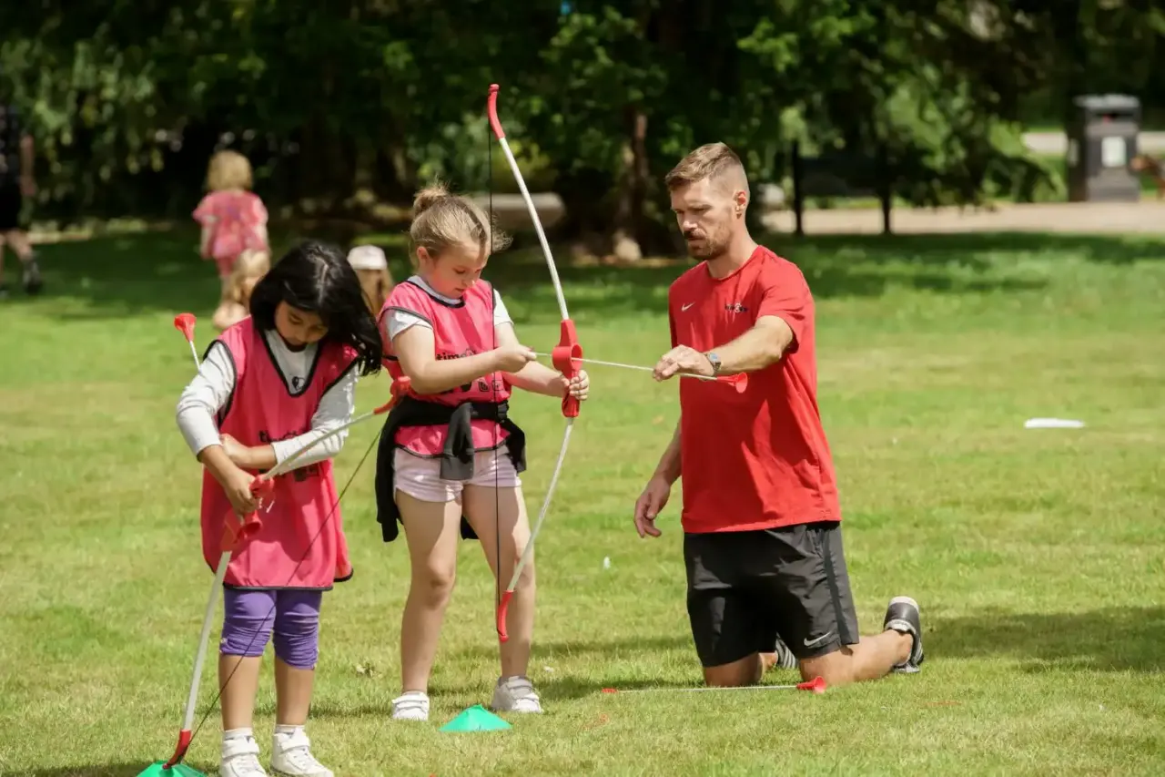An instructor teaches archery to two children on a sunny grass field, with other activity participants visible in the background.