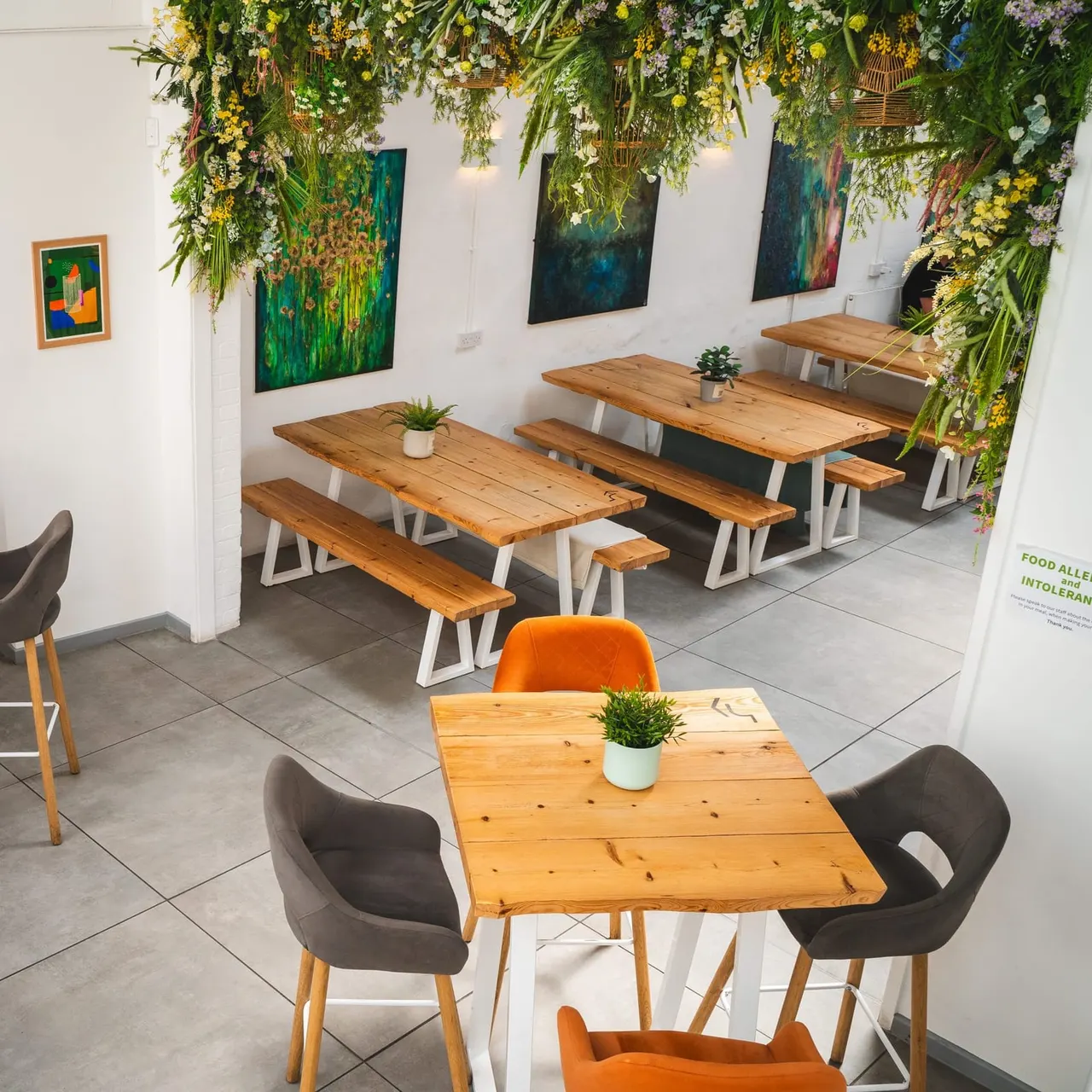 Interior of a casual dining venue with wooden tables, coloured chairs, hanging floral installations and artwork on white walls.