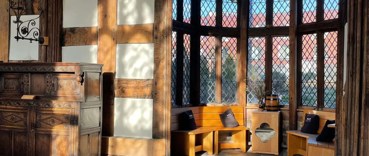 Historic interior with wooden furniture, latticed windows showing sunset sky, and period architectural details including carved wooden cabinet and decorative ironwork.