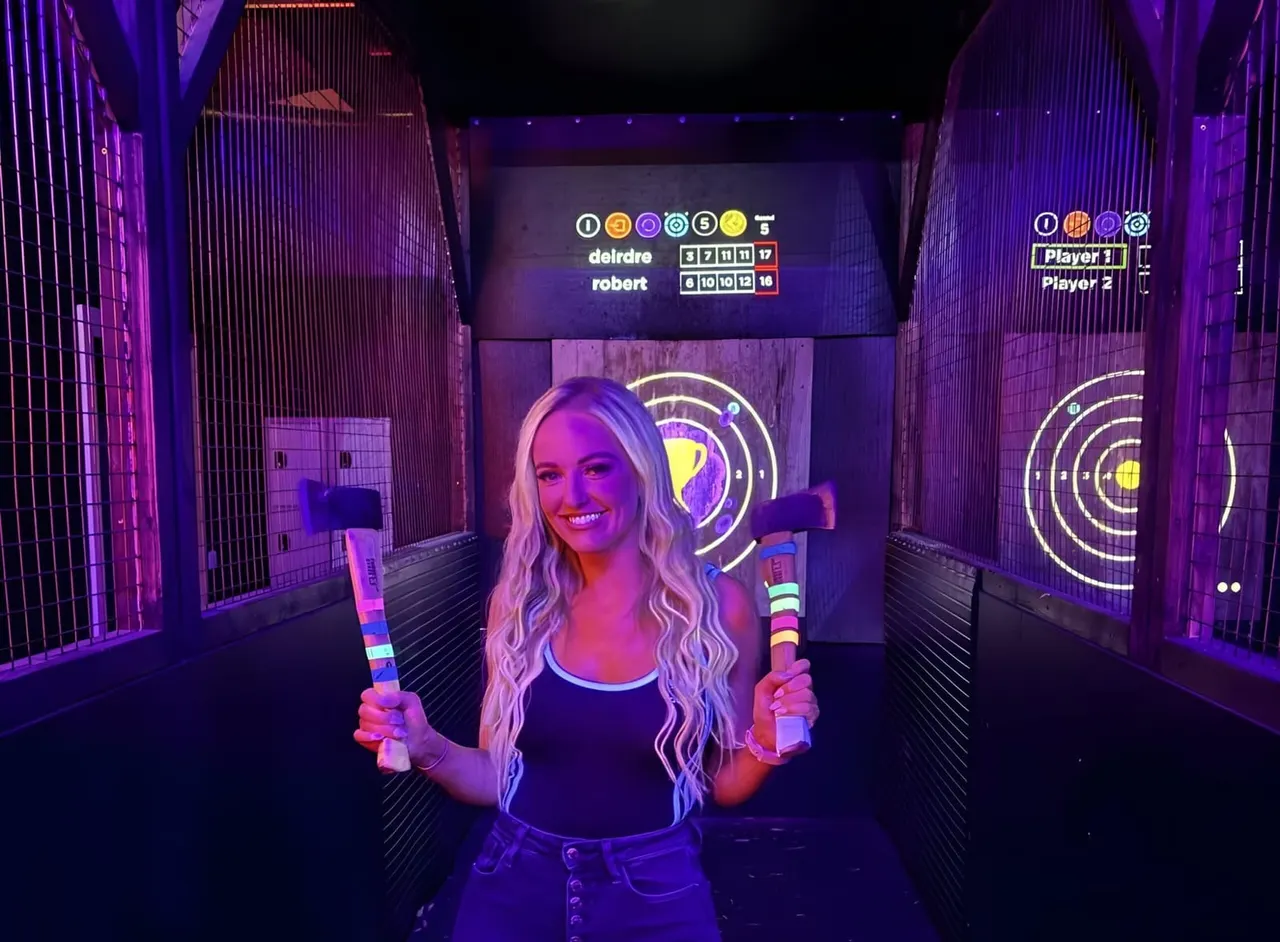 A woman holding neon-lit foam bats smiles at the camera in a neon-lit arcade game booth with circular target displays.