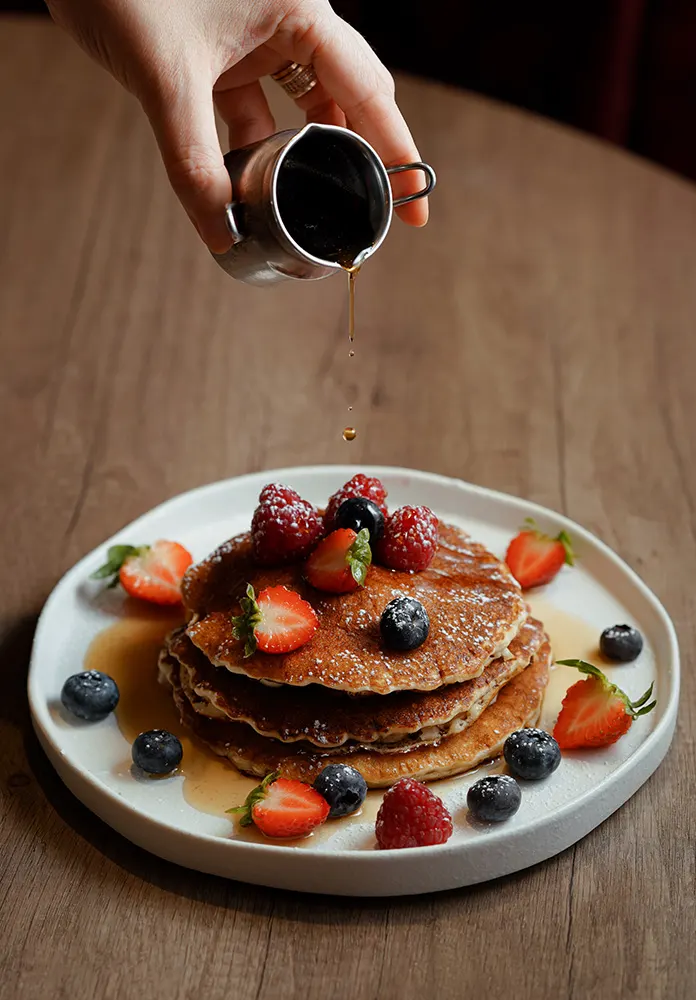 Hand pouring syrup over a stack of pancakes topped with fresh berries on a wooden table.