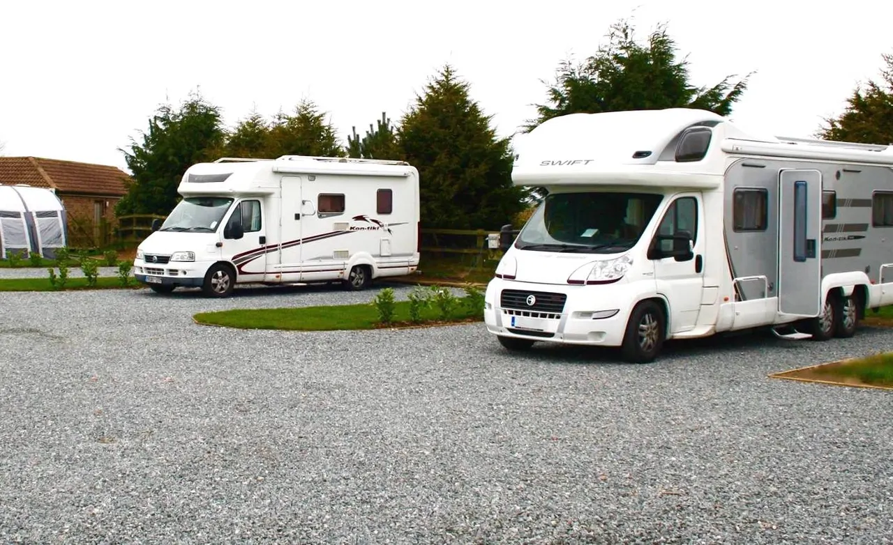 Three white motorhomes parked on gravel at a campsite with trees and a cottage building in background.
