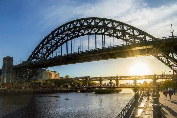 The Tyne Bridge's distinctive arched steel structure spans above the river at sunset, with people walking along the riverside path below.