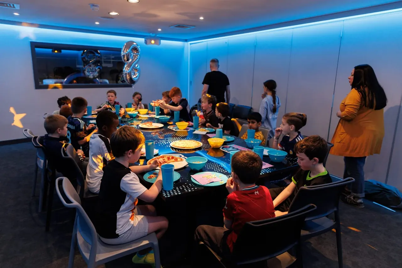 Group of children eating pizza and food at a table during a party in a blue-lit indoor venue.
