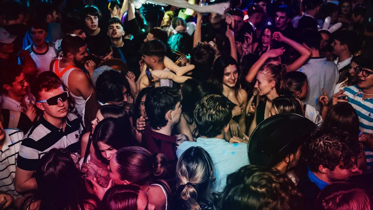 Large crowd of people dancing and celebrating in a nightclub with colourful neon lighting.