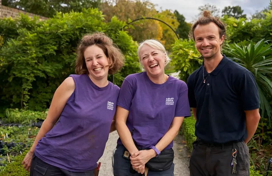 Three smiling staff members in purple branded shirts stand on a garden path surrounded by lush greenery and shrubs.