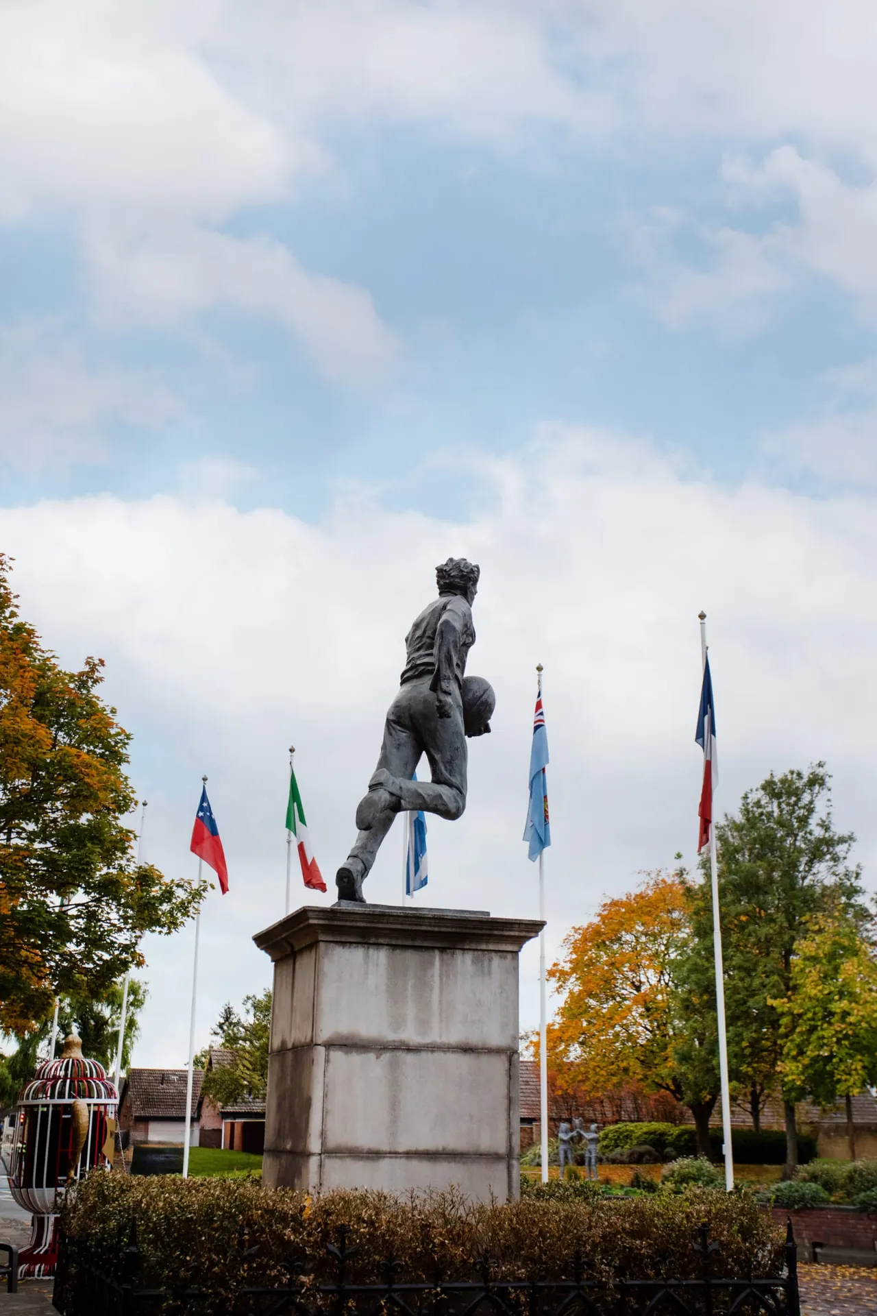 Stone statue of a rugby player in mid-stride on a plinth, surrounded by international flags and autumn trees.