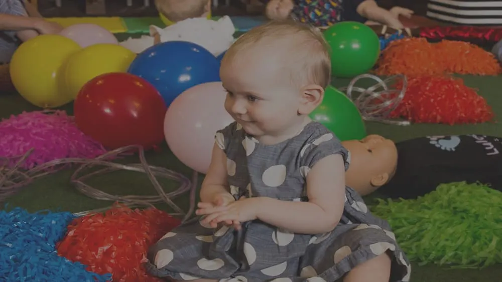 A toddler sits on a soft play mat surrounded by colorful balloons and textured sensory toys.