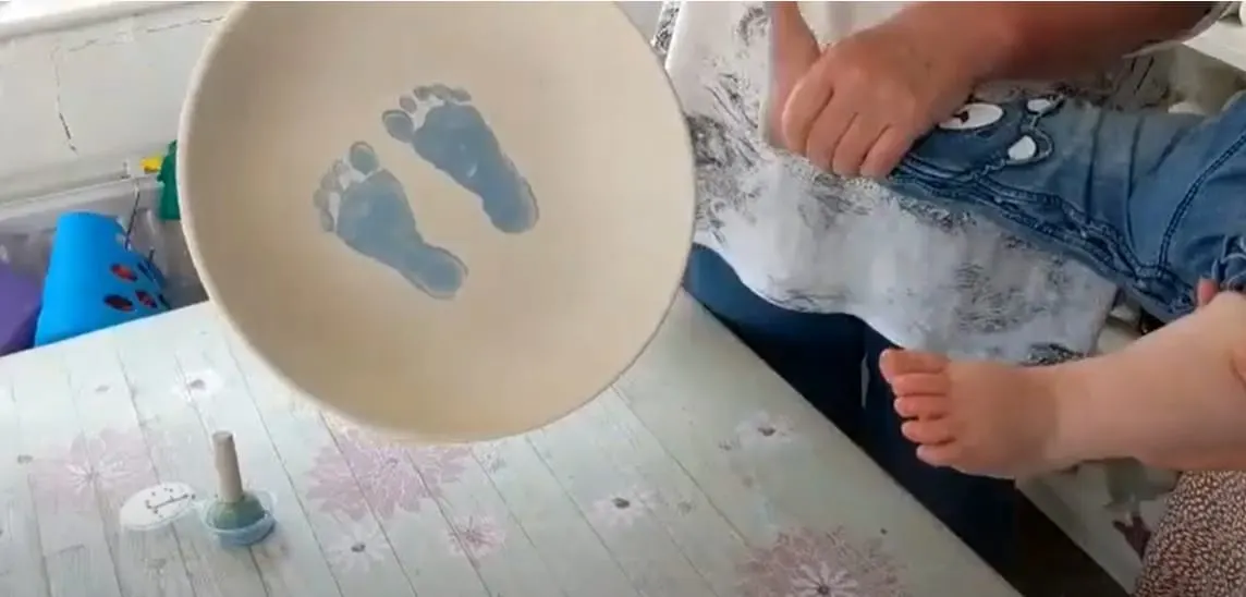 A young child's bare feet leave blue footprints in a shallow bowl of water during a messy play activity.
