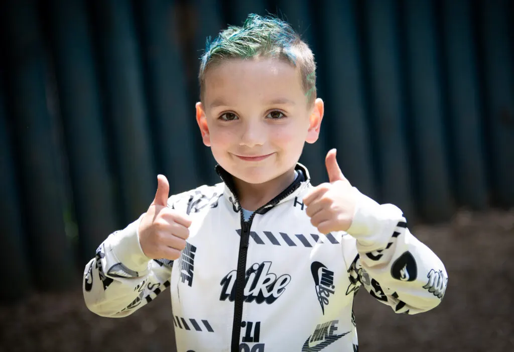 Young child in white and black sports outfit giving double thumbs up to camera outdoors.