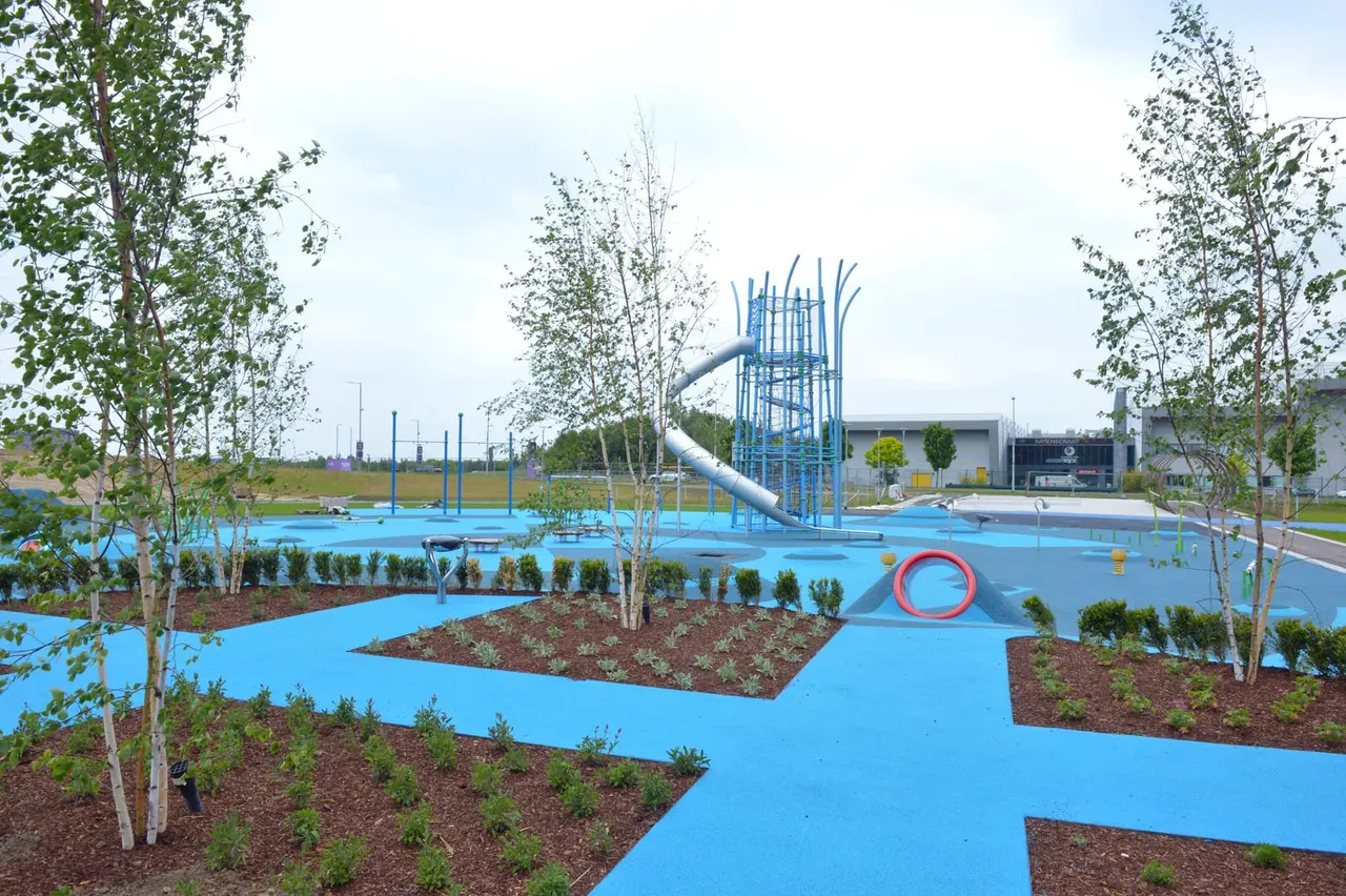 Modern adventure playground with blue surfacing, climbing structures, slide tower and tunnel features surrounded by newly planted trees.