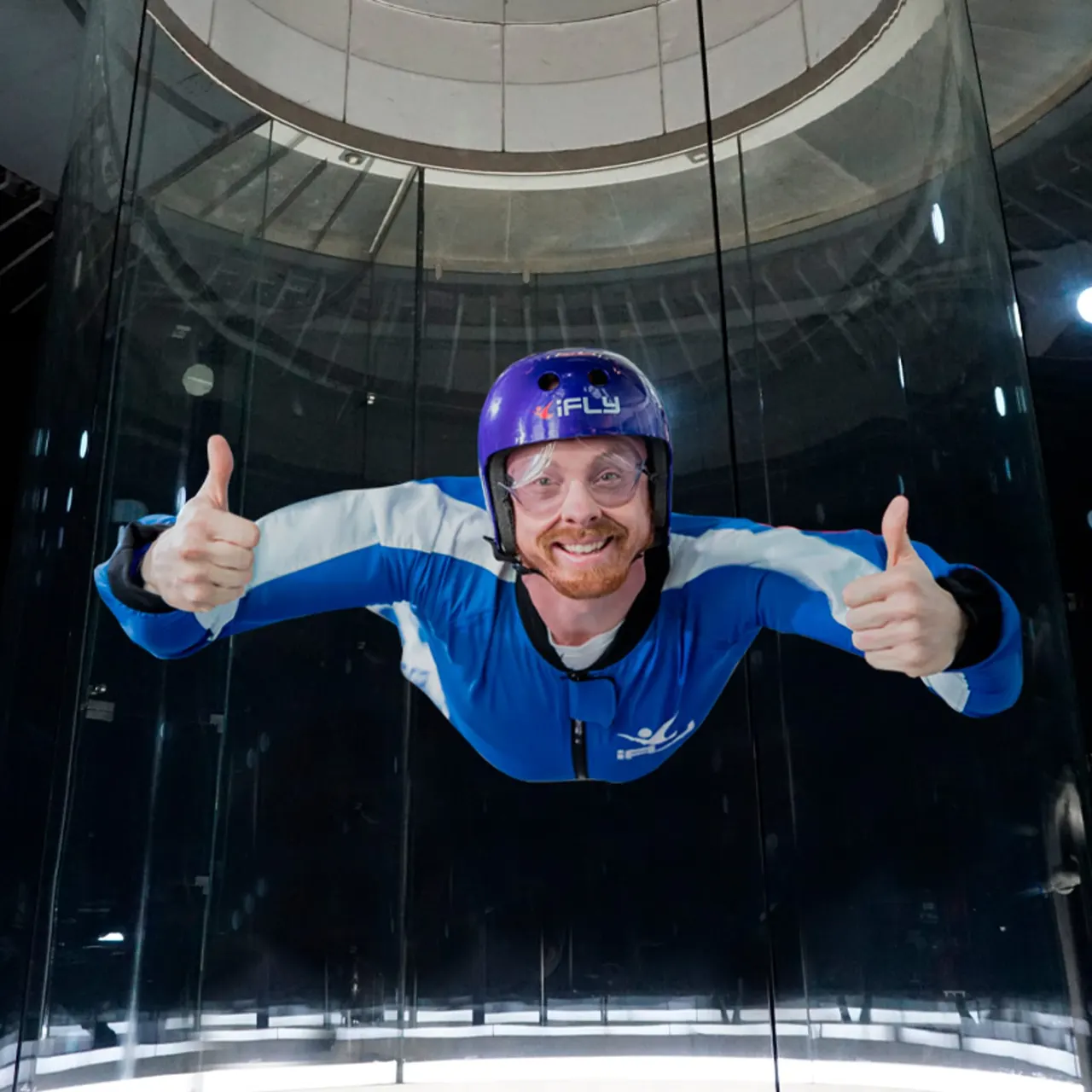 Man in blue flight suit and helmet giving double thumbs up whilst indoor skydiving in a wind tunnel.
