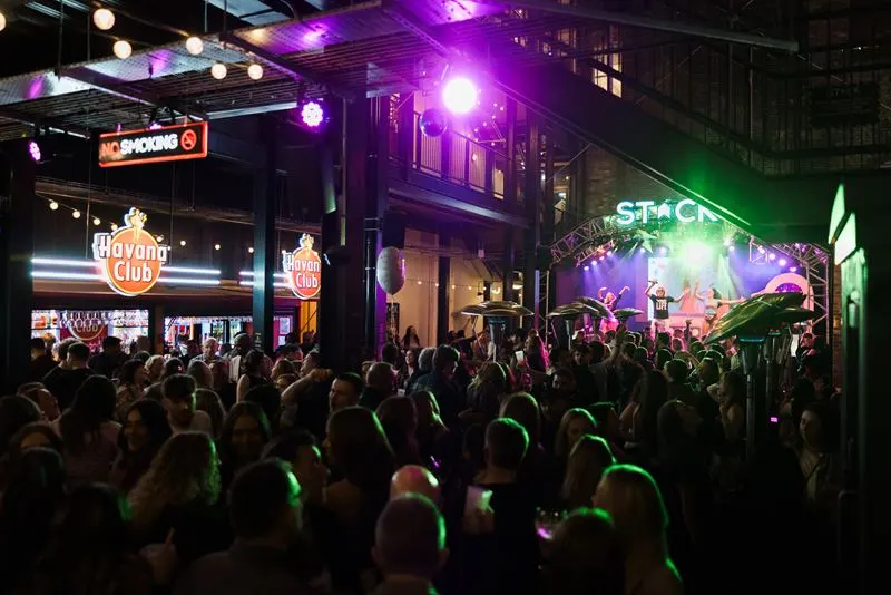 Large crowd of people dancing and watching a live band perform on an outdoor stage at night with purple and green stage lighting.