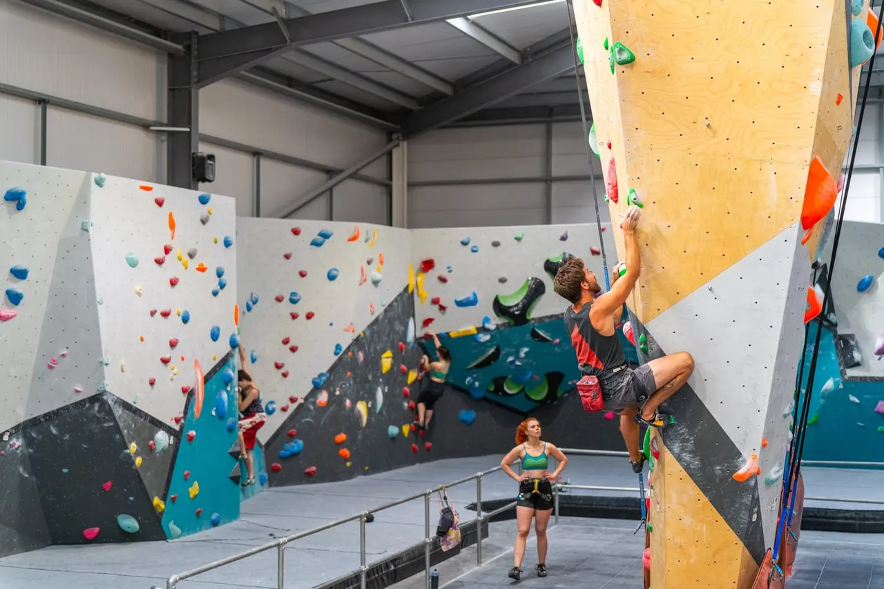 Indoor climbing wall facility with multiple climbers of varying ages ascending walls with coloured holds whilst others watch from below.