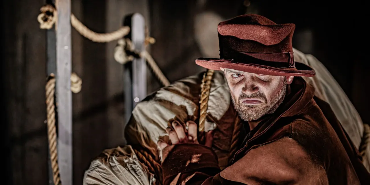 Man in period costume holding rope inside a dark dungeon setting.