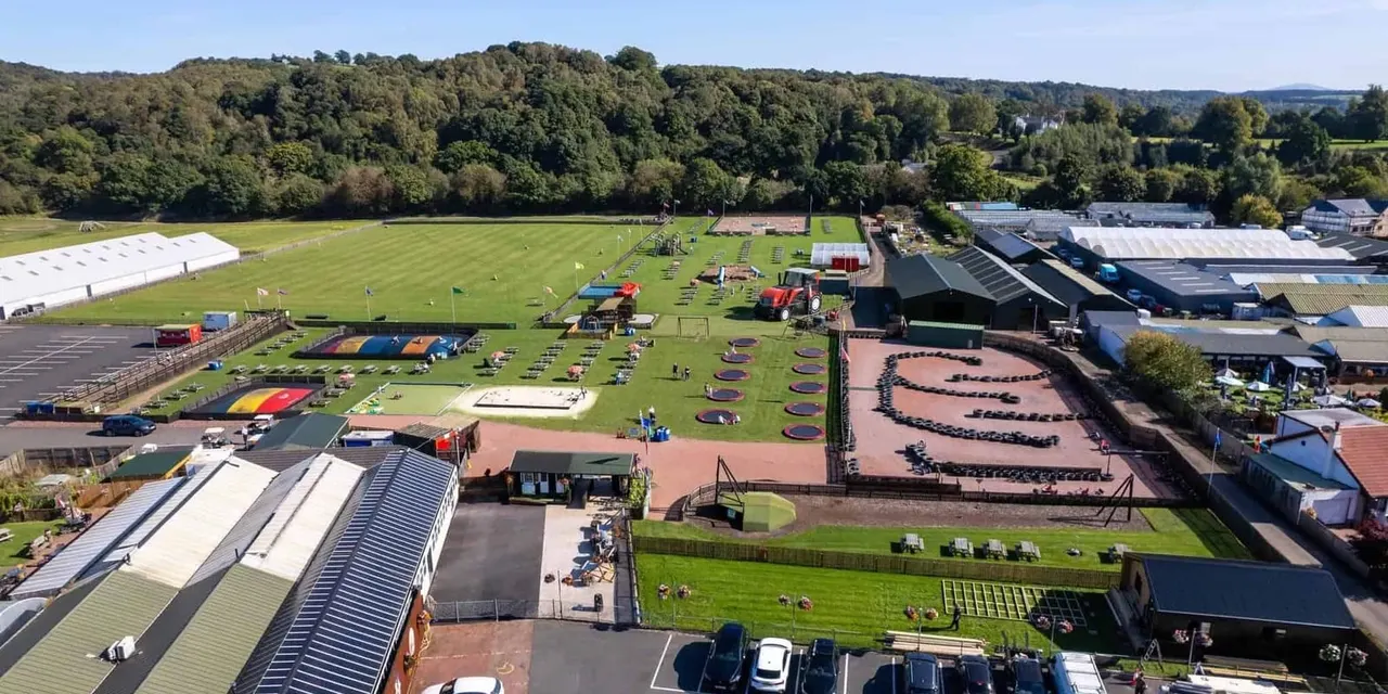 Aerial view of a large leisure park with activity areas, trampolines, play equipment, green fields and woodland backdrop.